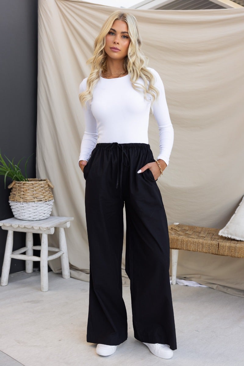 Stylish model wearing black Peru pants with elastic waistband and wide legs, paired with a white long-sleeve top.