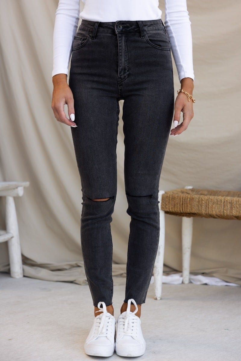 Washed black denim skinny jeans with mid-high waist and distressed rips, styled with white top and sneakers.