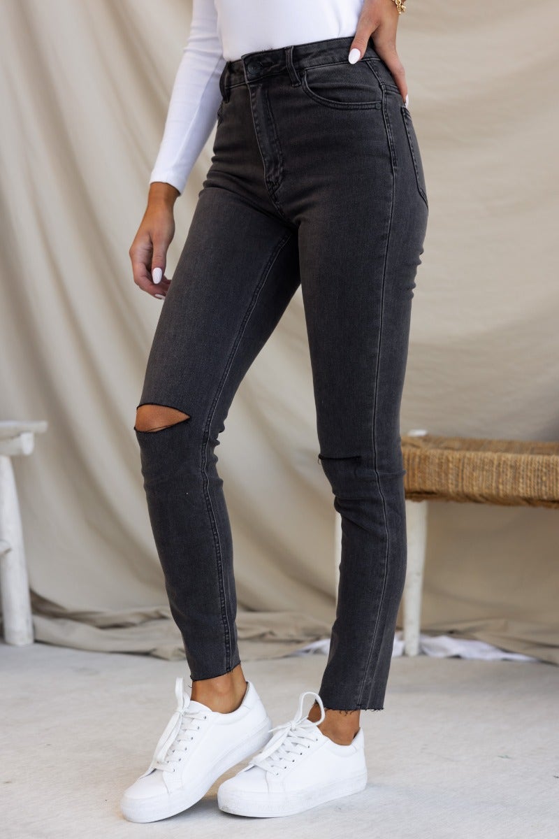 Dion Jeans in washed black denim with mid-high waist, distressed rips, raw hemline, and stretchy fit.