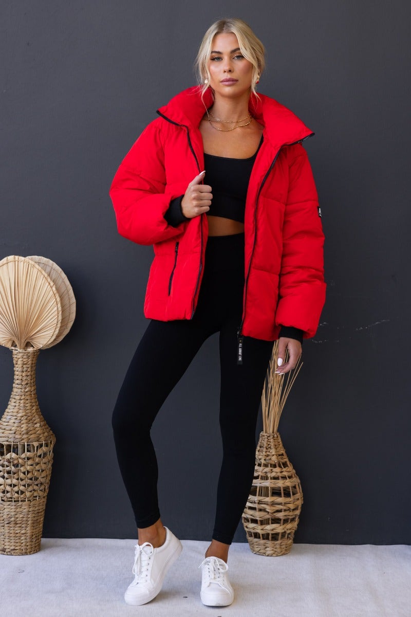 Delila Puffer Jacket in vibrant red, featuring high neckline, removable hood, and zip-up front, styled with black leggings.