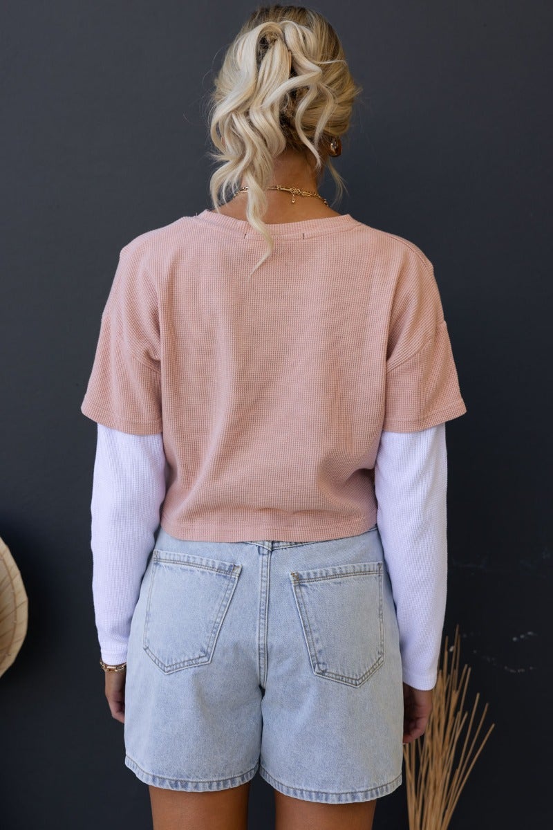 Model wearing a relaxed crop Tyrra Top in textured pink with white sleeves, paired with denim shorts, showcasing back view.