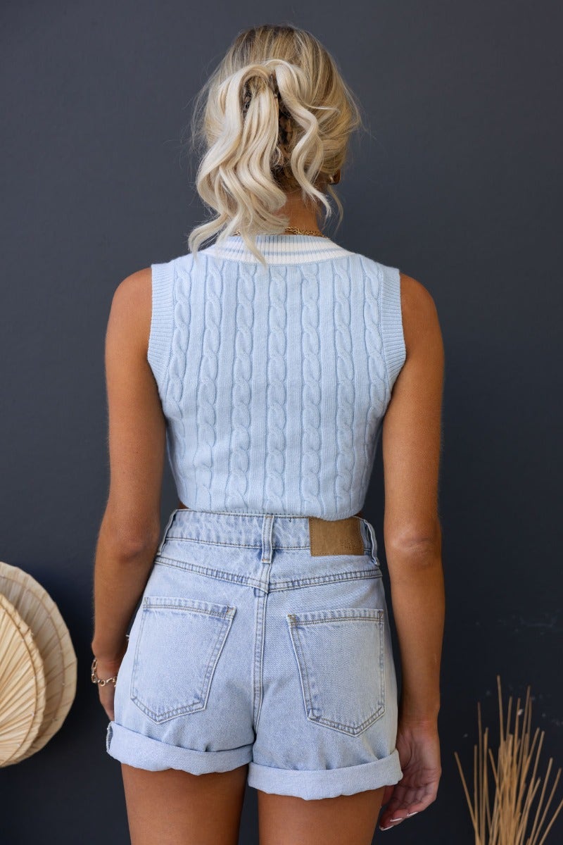 Back view of a woman in a light blue Beia Top knit vest, paired with high-waisted denim shorts against a dark background.