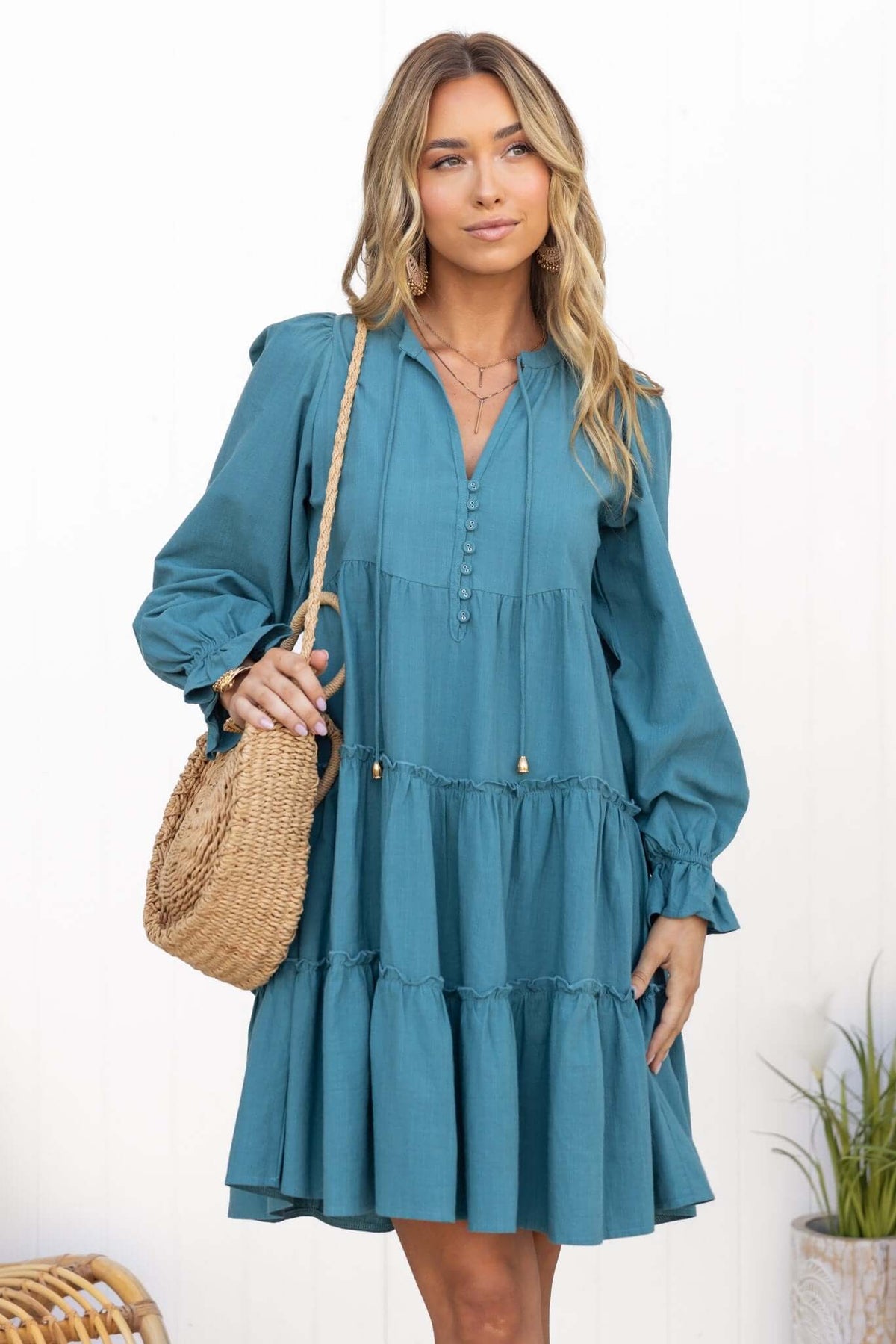 Model wearing Boone Dress in teal with V-neckline, tie detail, and tiered ruffle skirt, styled with a straw bag.