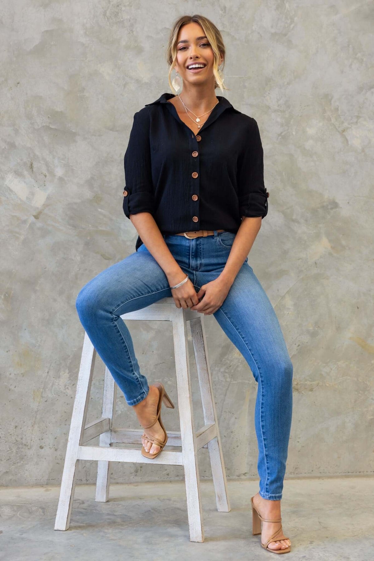 Model wearing a black Muscat shirt with button-up front, paired with blue jeans and heels, sitting on a stool.