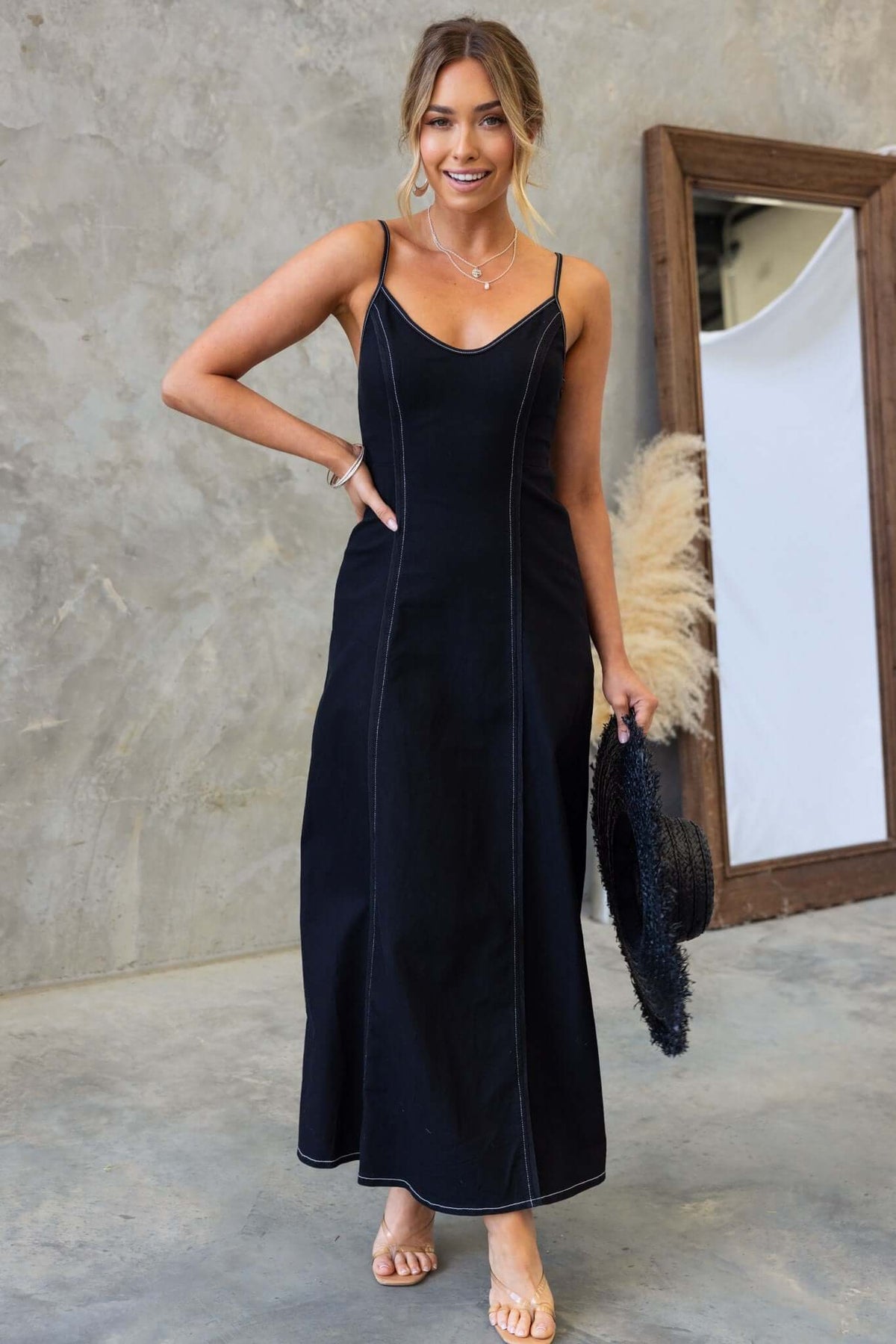 Model wearing a black denim maxi dress with adjustable straps and side zip closure, styled with a hat.
