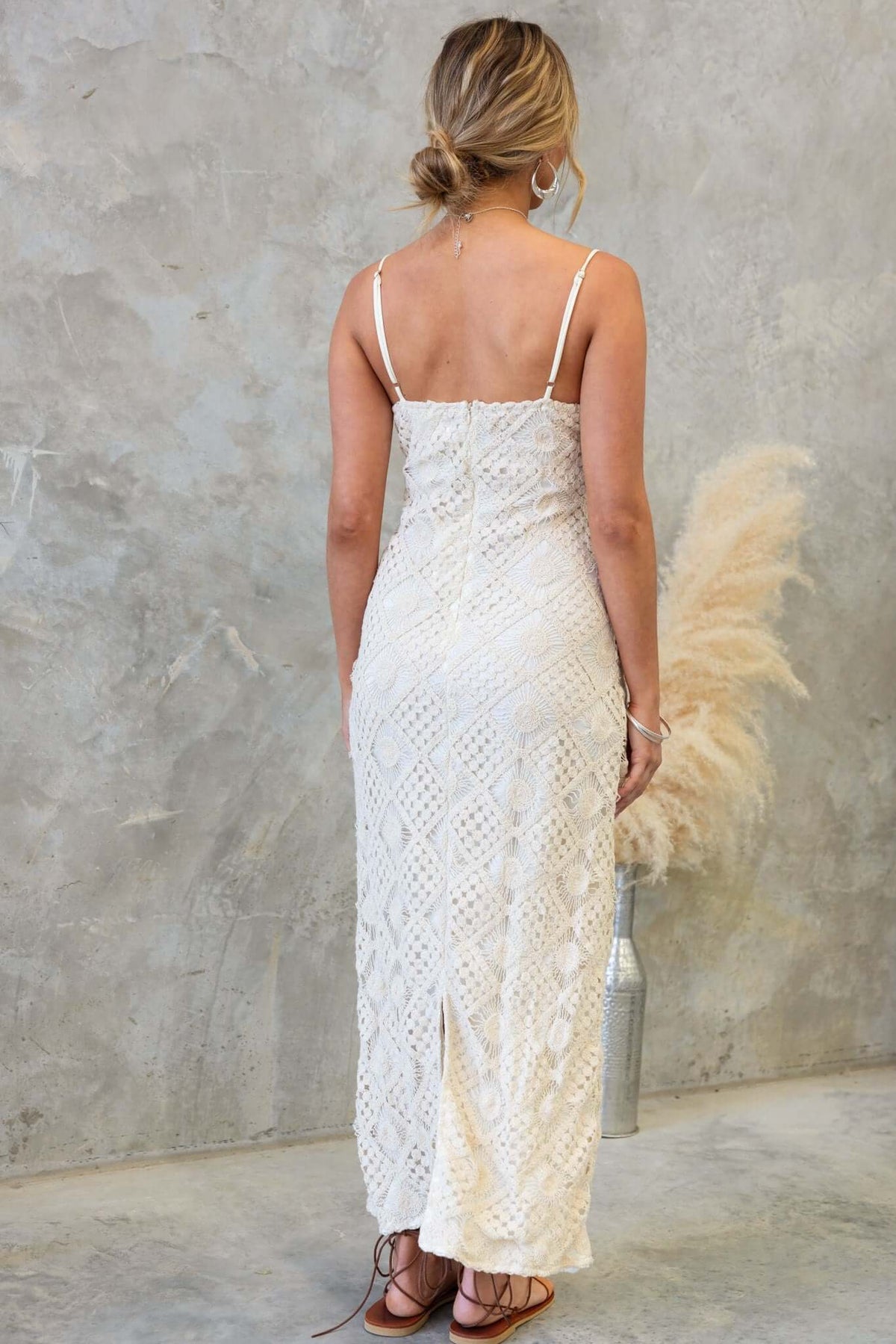 Back view of Udele Maxi Dress in crochet material with adjustable straps and a back slit, styled with a rustic background.