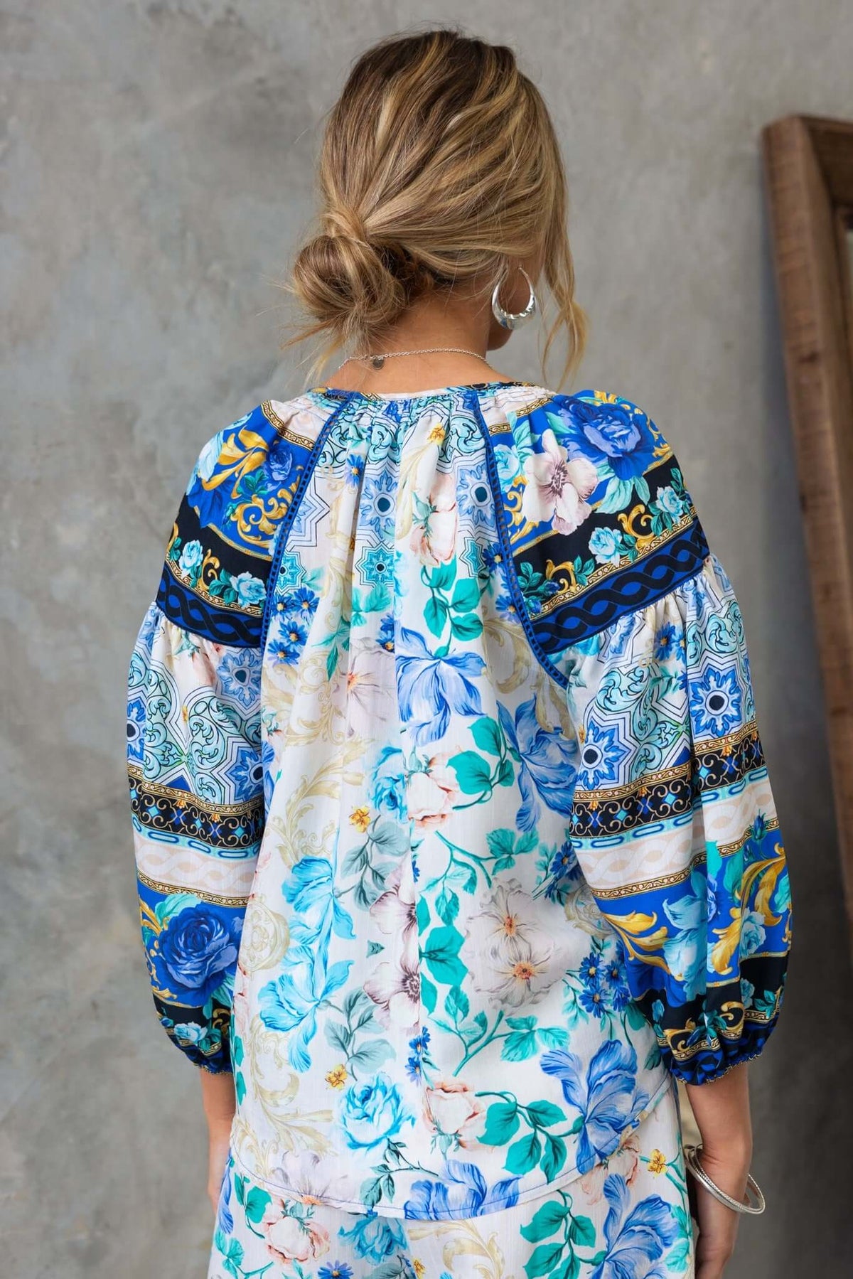 Back view of a woman in a floral print top with puffed sleeves and vibrant blue patterns against a textured background.