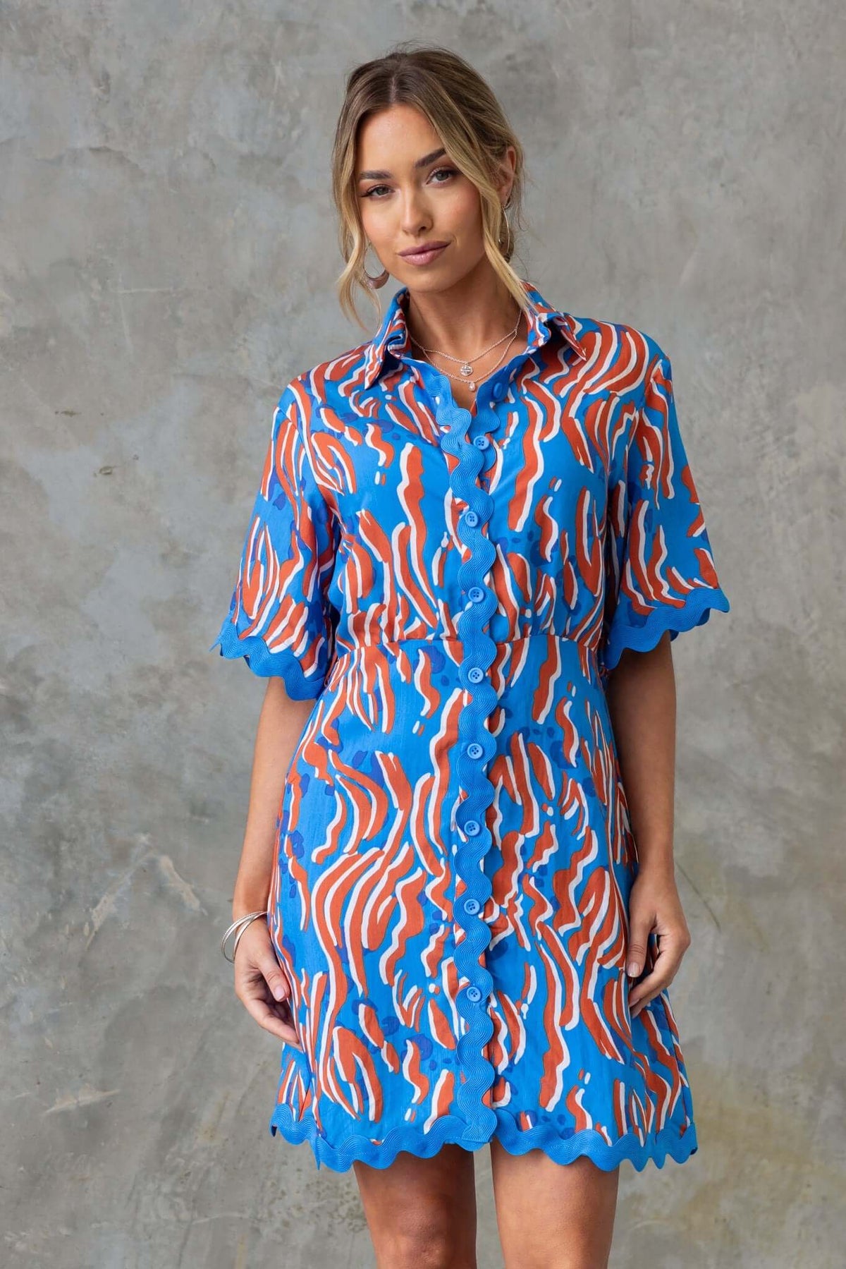Model wearing the Alfonzo Dress featuring a vibrant blue and orange pattern, button-up front, and scalloped edges.
