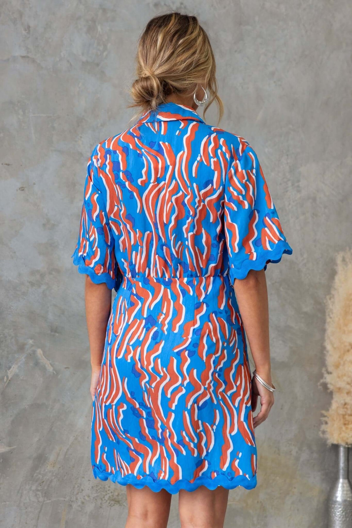 Back view of the Alfonzo Dress featuring vibrant blue, orange, and white wavy patterns, with a scalloped hem and fitted waist.