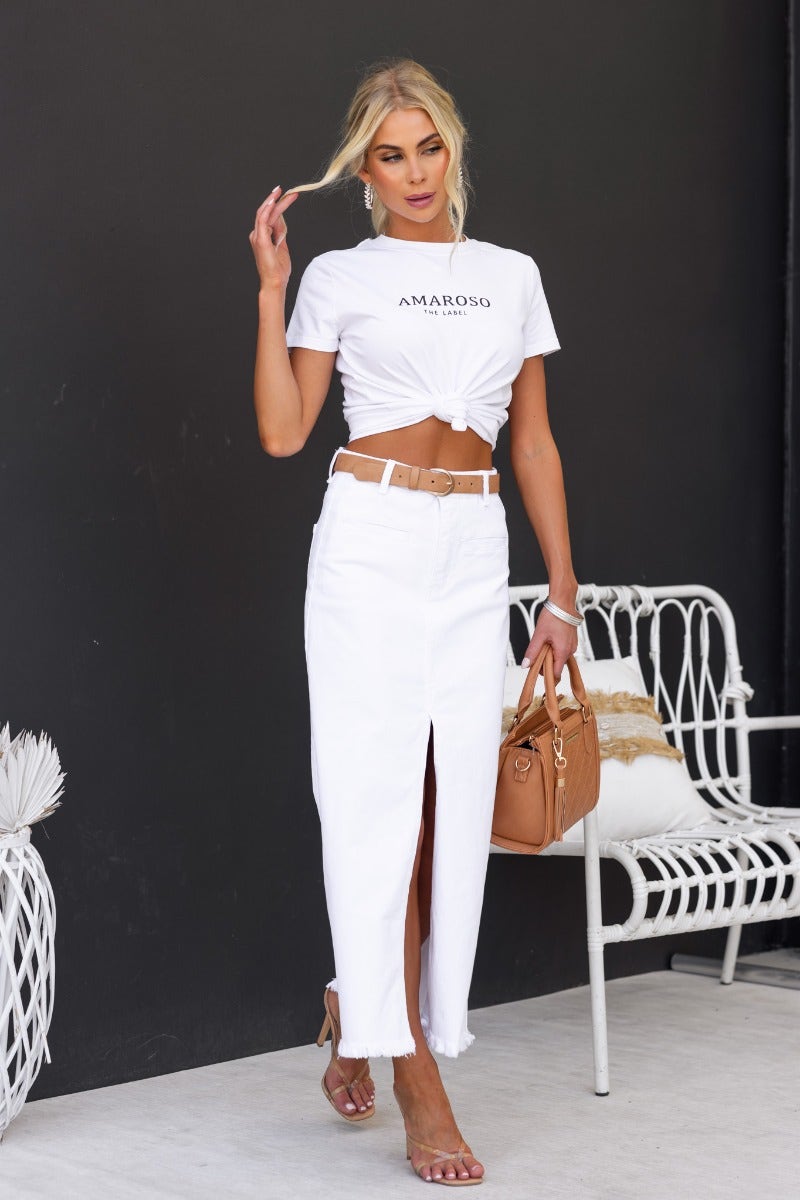 Woman in a white denim maxi skirt and cropped tee, posing with a handbag in a stylish setting.