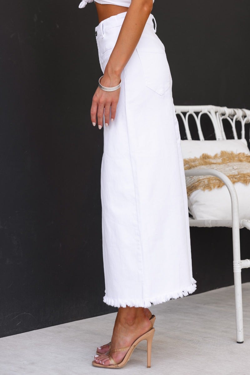 Side view of a white denim maxi skirt with frayed hemline and pockets, styled with heels and sitting on a chair.