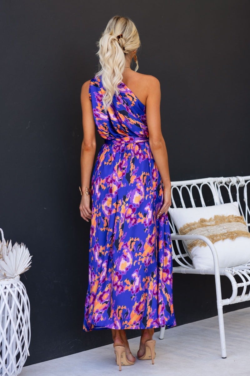 Back view of a woman in a colorful one-shoulder maxi dress with an elastic waistband and removable belt.