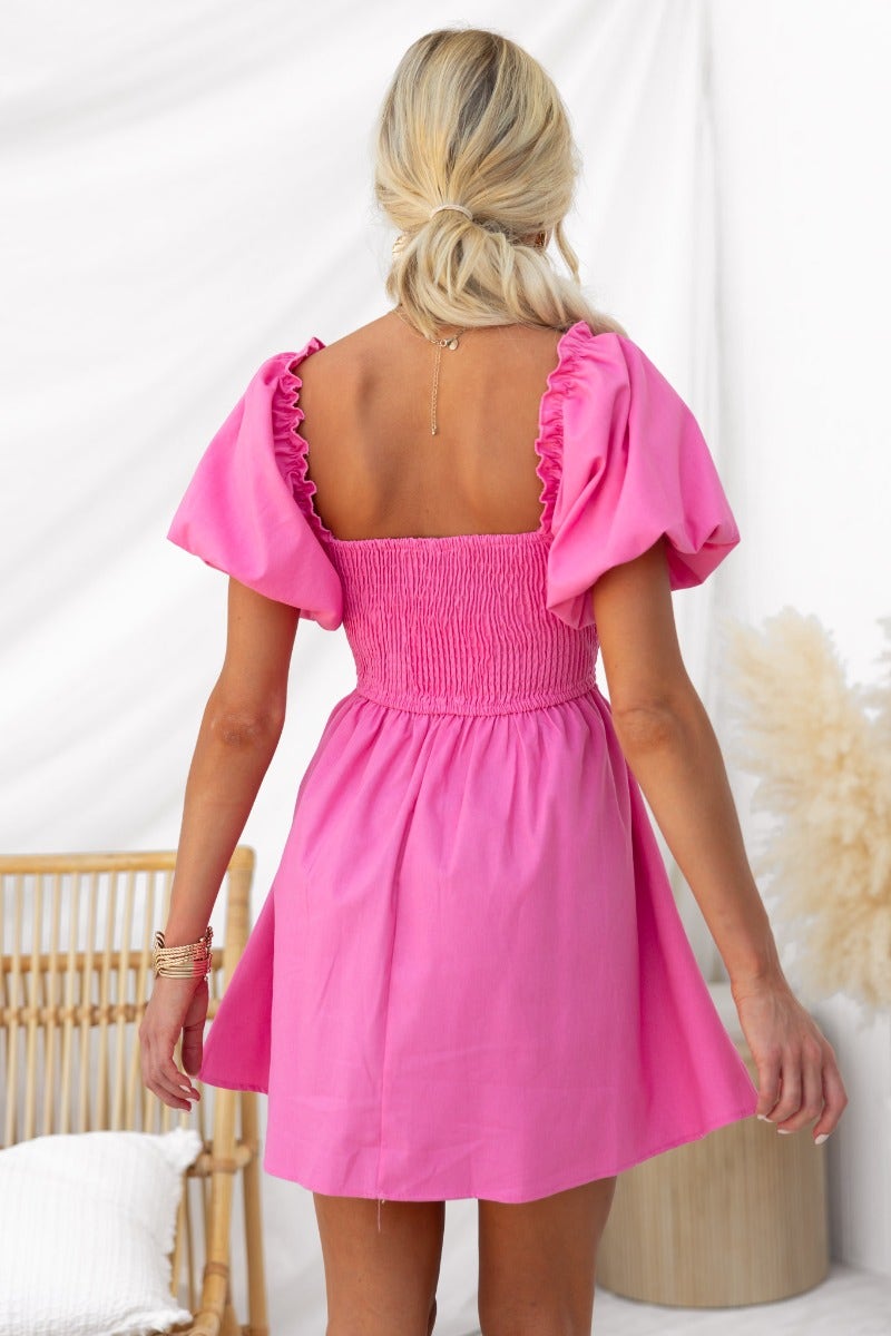 Back view of a woman wearing a pink smocked dress with puff sleeves, styled in a modern indoor setting.