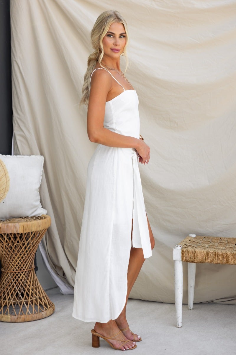 Model wearing a white midi dress with adjustable straps and a tie skirt, styled in a chic setting.