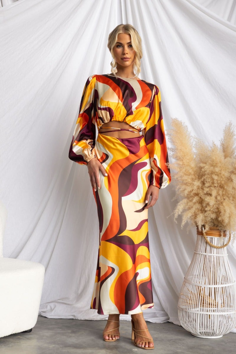 Model wearing a colorful satin Goldie maxi dress with cut-outs, puff sleeves, and a tie detail, styled with a minimal backdrop.