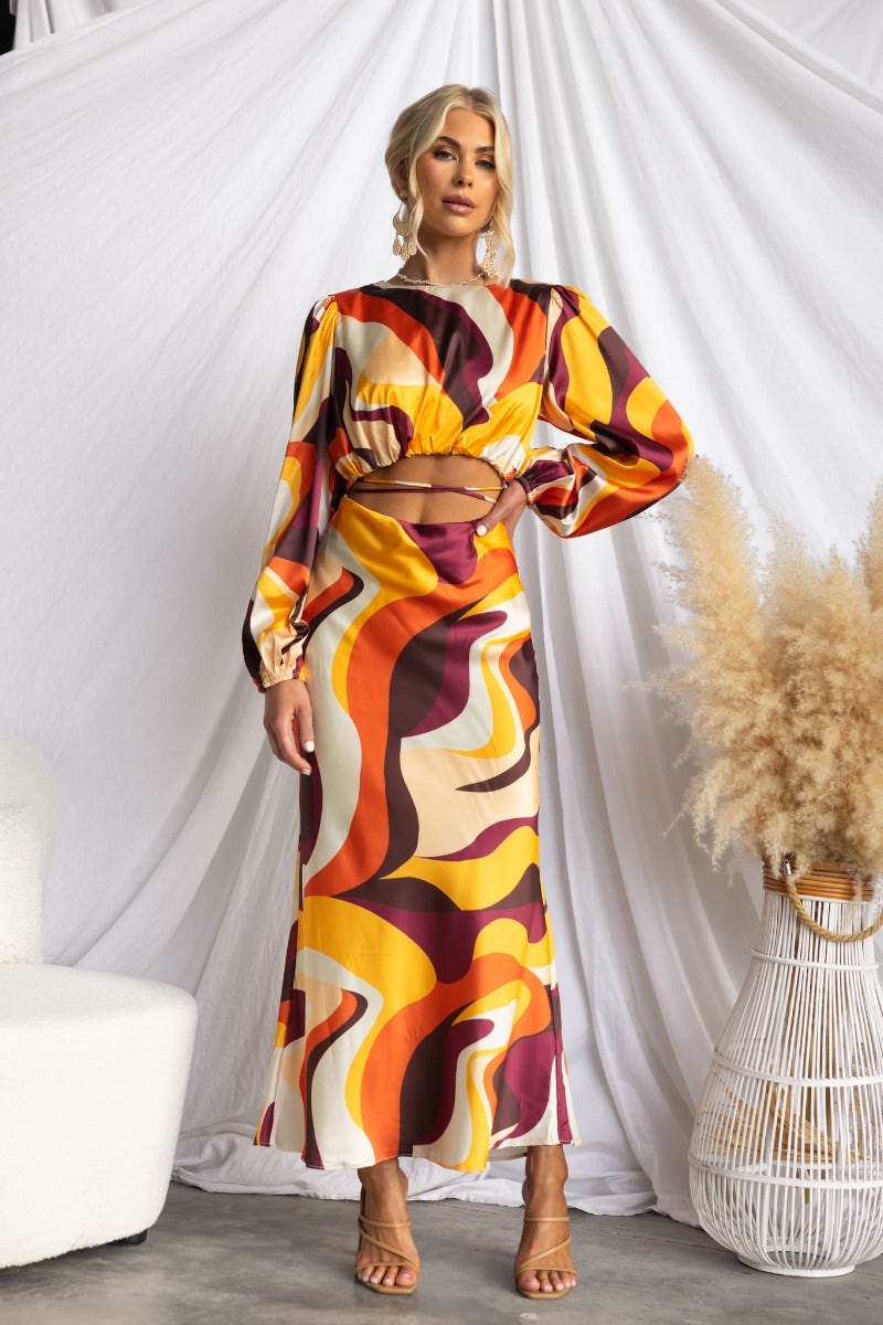 Model wearing a colorful satin Goldie maxi dress with cut-out waist and elastic sleeves, styled in a chic indoor setting.