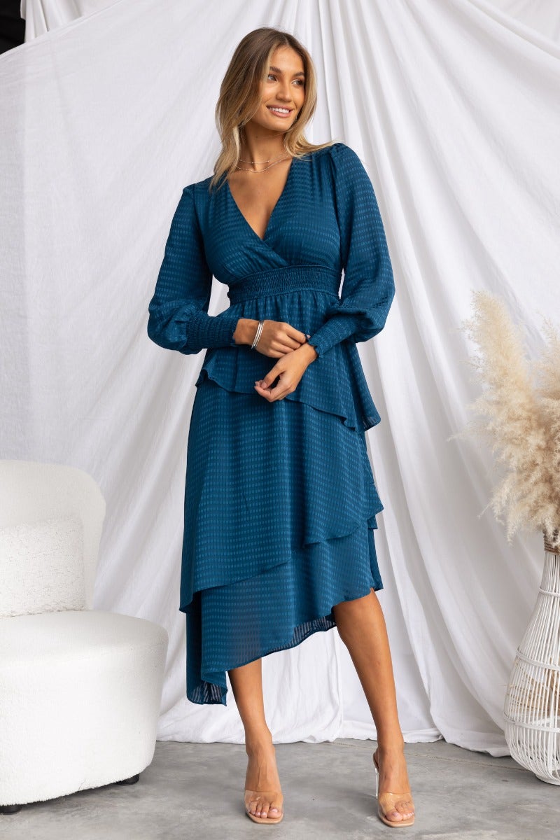 Briena Midi Dress in teal with V-neckline, multi-tier ruffle skirt, and asymmetrical hemline, styled in a chic indoor setting.
