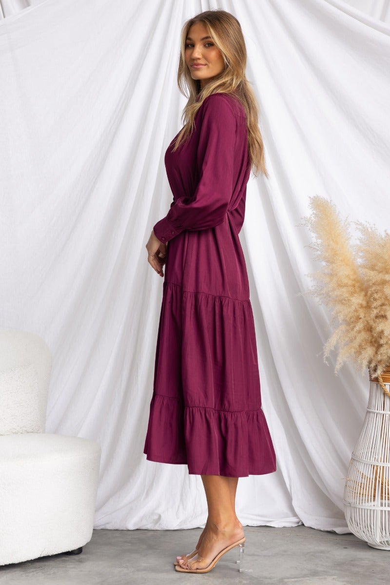 Side view of a model wearing a purple midi dress with a tiered skirt and long sleeves, styled for a relaxed fit.