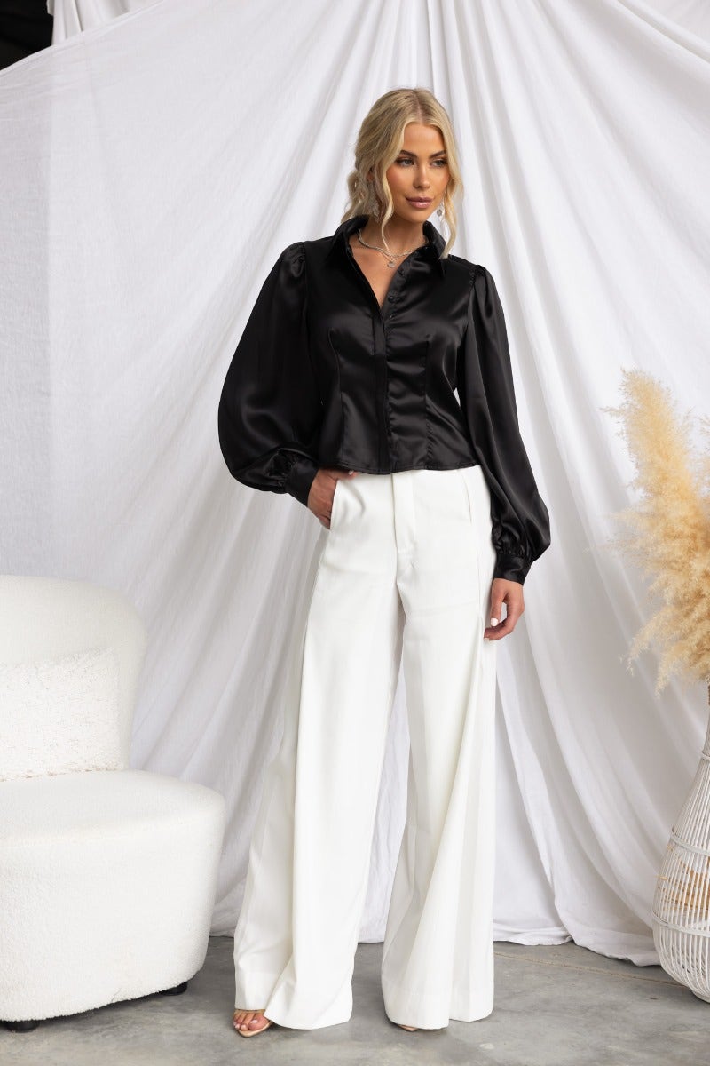 Model wearing the Iva Top, a black satin button-up top paired with white wide-leg pants against a soft backdrop.