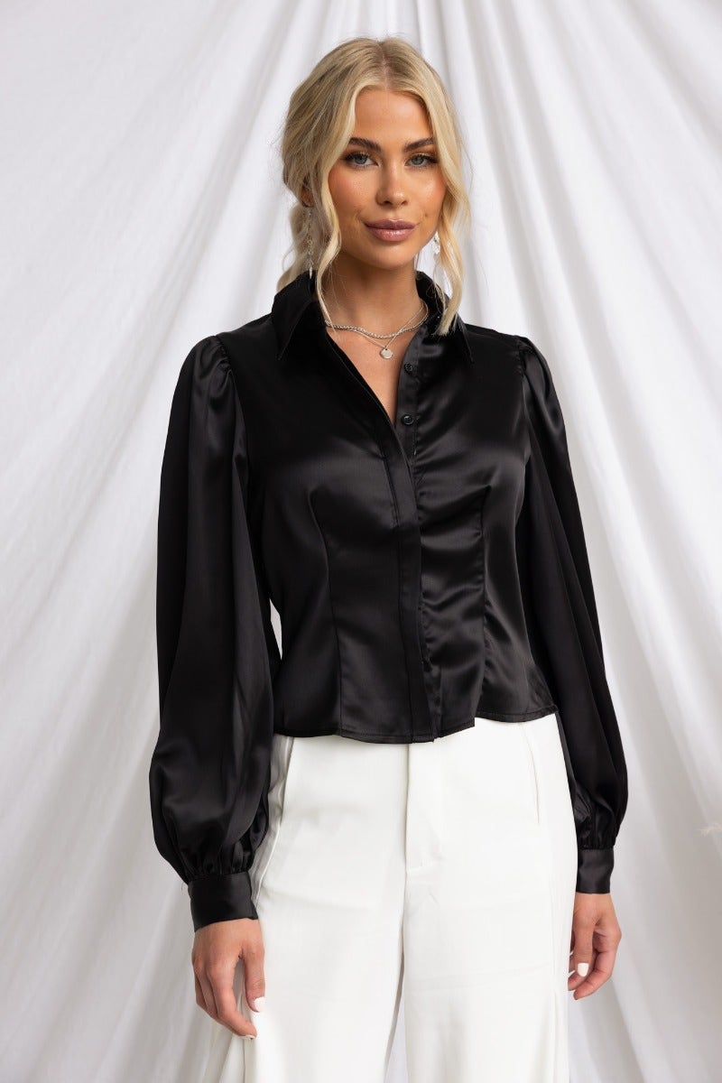 Model wearing a black satin Iva Top, featuring a collar and button-up front, styled with white trousers.