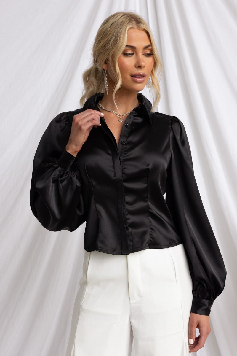 Model wearing the Iva Top in black satin with a collar and button-up front, styled with white pants and layered jewelry.