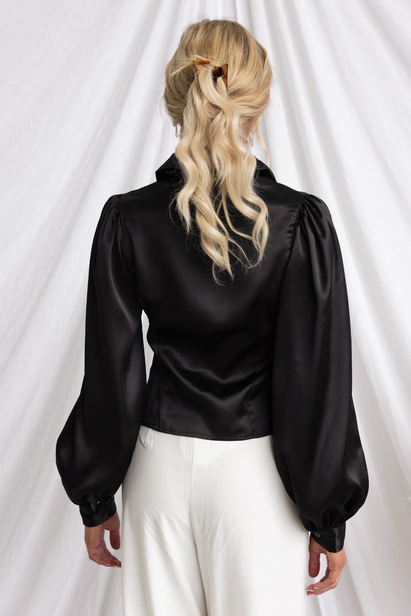 Back view of the Iva Top, a black satin button-up with puff sleeves, styled with white pants.