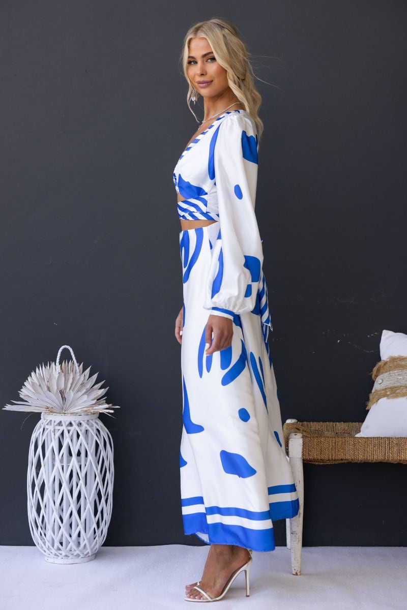 Model wearing Qasim Maxi Dress in blue and white with twist detail and cut-out at waist, styled with strappy heels.