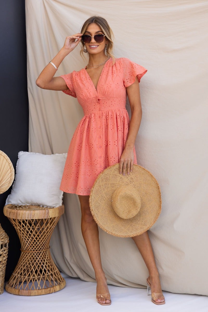 Model wearing a coral Udela Dress with V-neckline and button-up front, holding a straw hat in a stylish pose.