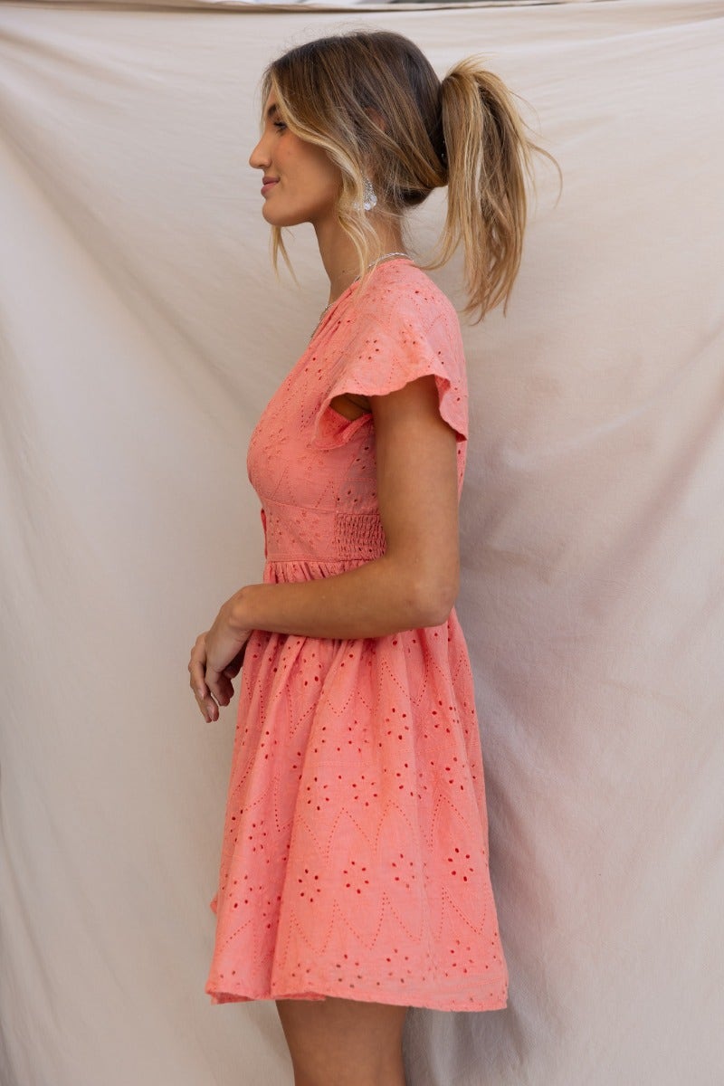 Profile view of a woman wearing a coral Udela Dress made of Anglaise material with a V-neckline and flutter sleeves.