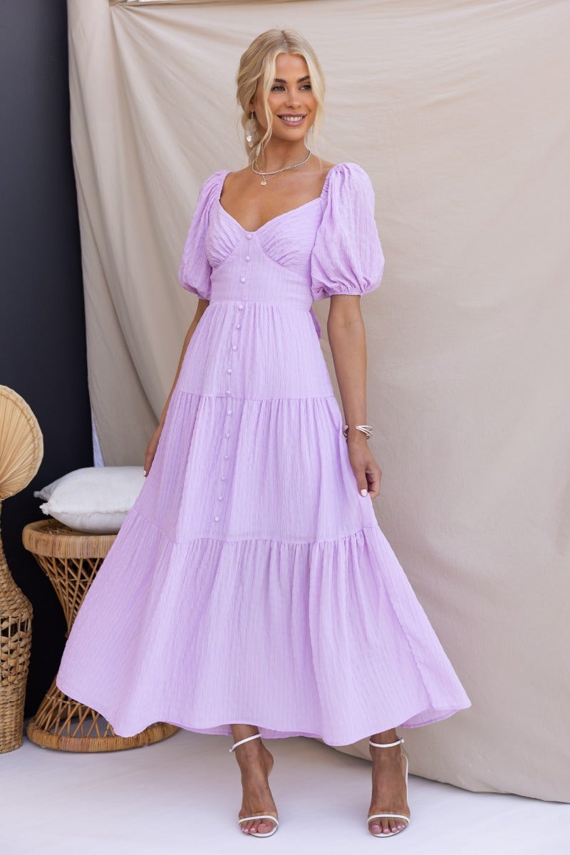 Model wearing Nammos Maxi Dress in light purple, featuring puff sleeves and button details, set against a neutral backdrop.