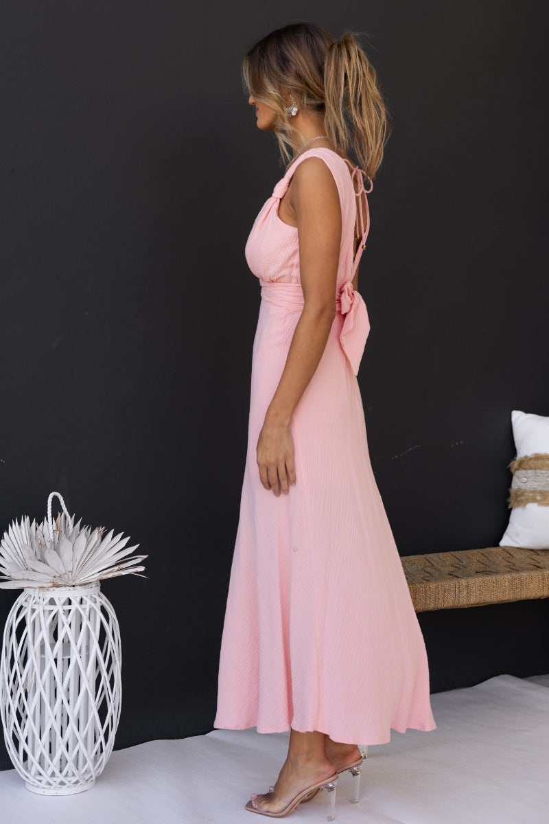 Side view of a model wearing the Gia Maxi Dress in pink with a tie detail and textured fabric, showcasing a flowing silhouette.