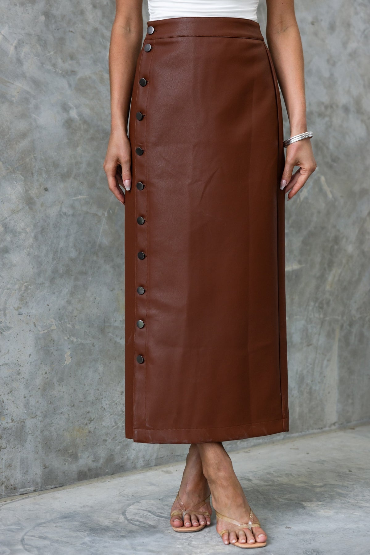 Brown faux leather maxi skirt with button-up front, styled with casual sandals, on a textured grey background.