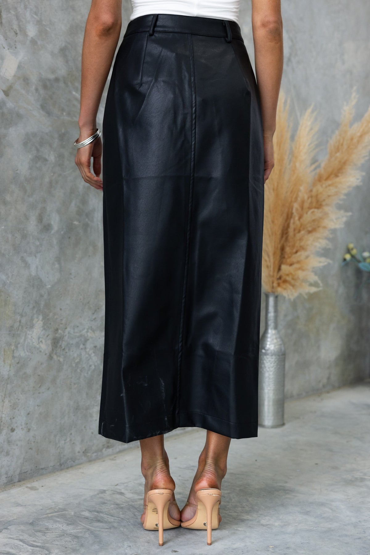 Rear view of black faux leather maxi skirt with mid-high waist fit, styled with heels in a modern setting.