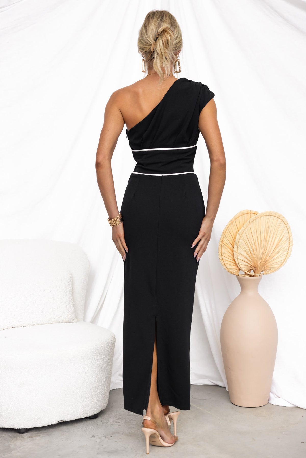 Back view of a woman in a black one-shoulder maxi dress with a split, styled with elegant accessories.