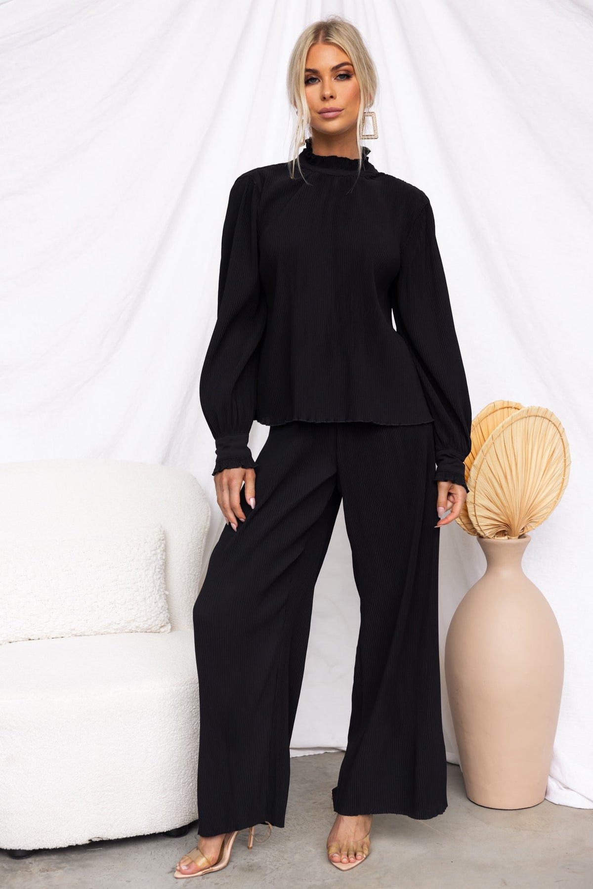 Black Barbara Top with high neckline and pleat design, styled elegantly with wide leg pants and a minimalist decor.