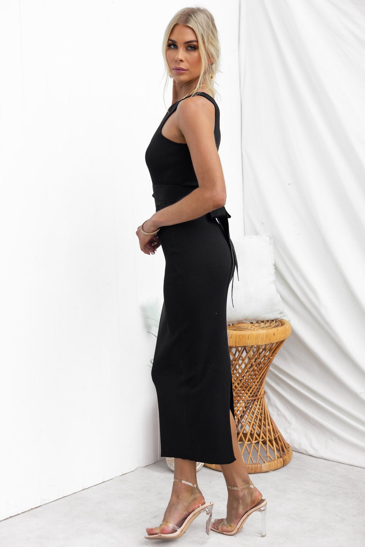 Side view of a woman in a black Lauryn Midi Dress with a round neckline and tie detail, styled with clear heels.