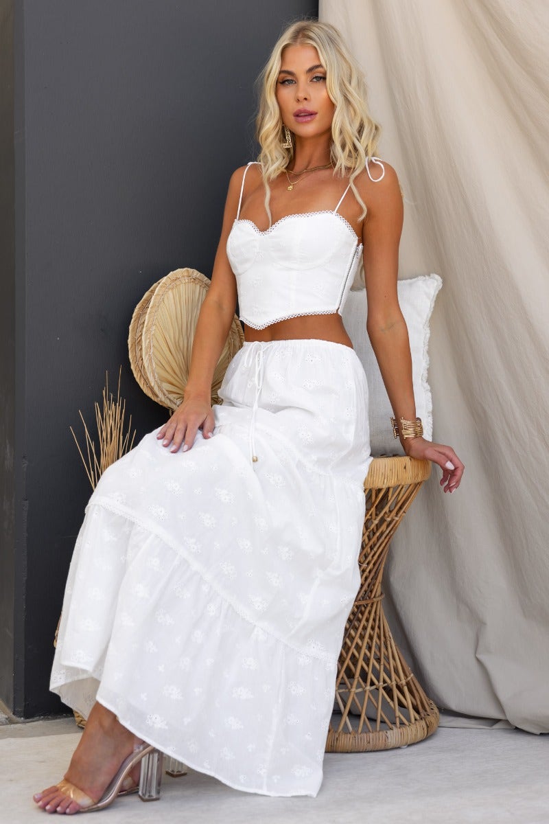 Model wearing a white Anglaise maxi skirt with an elastic waistband and a matching crop top, sitting on a chair.