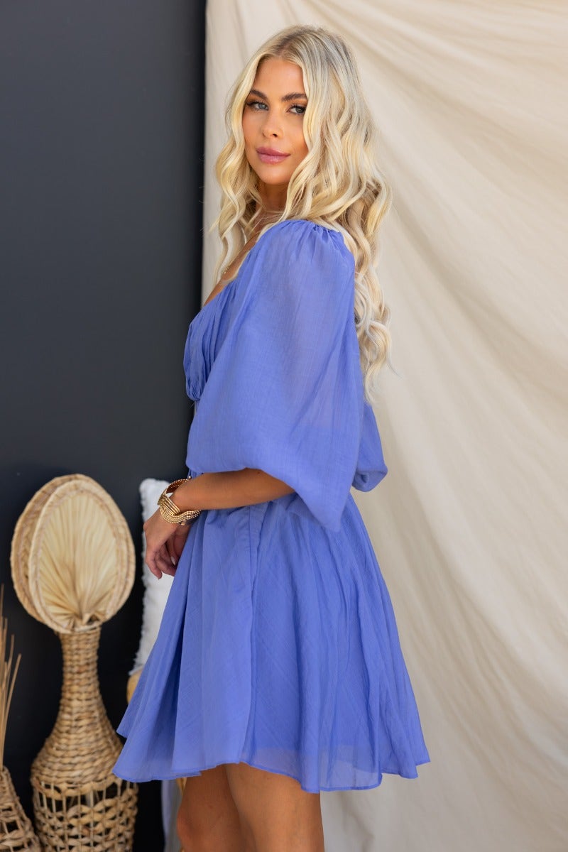 Model wearing a violet mini dress with elastic sleeves, back cut-out details, and flowing fabric.