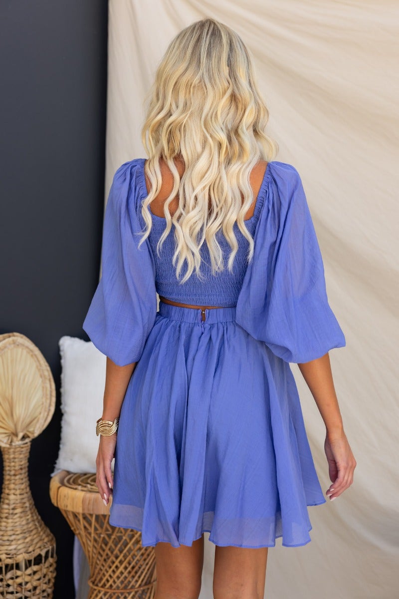 Back view of a woman in a violet mini dress with puff sleeves and a cut-out design, styled for summer fashion.