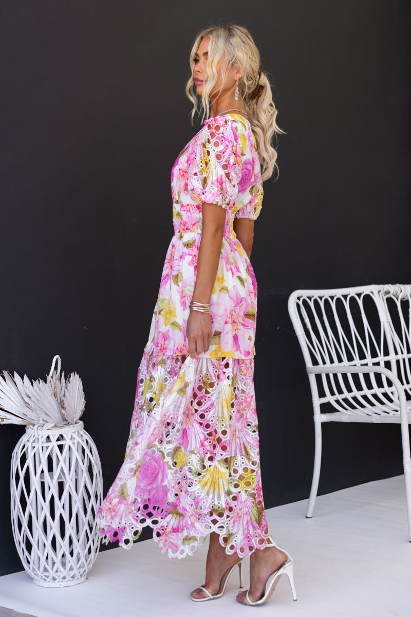 Woman in pink floral print maxi dress with V-neckline, standing next to a white wicker chair.