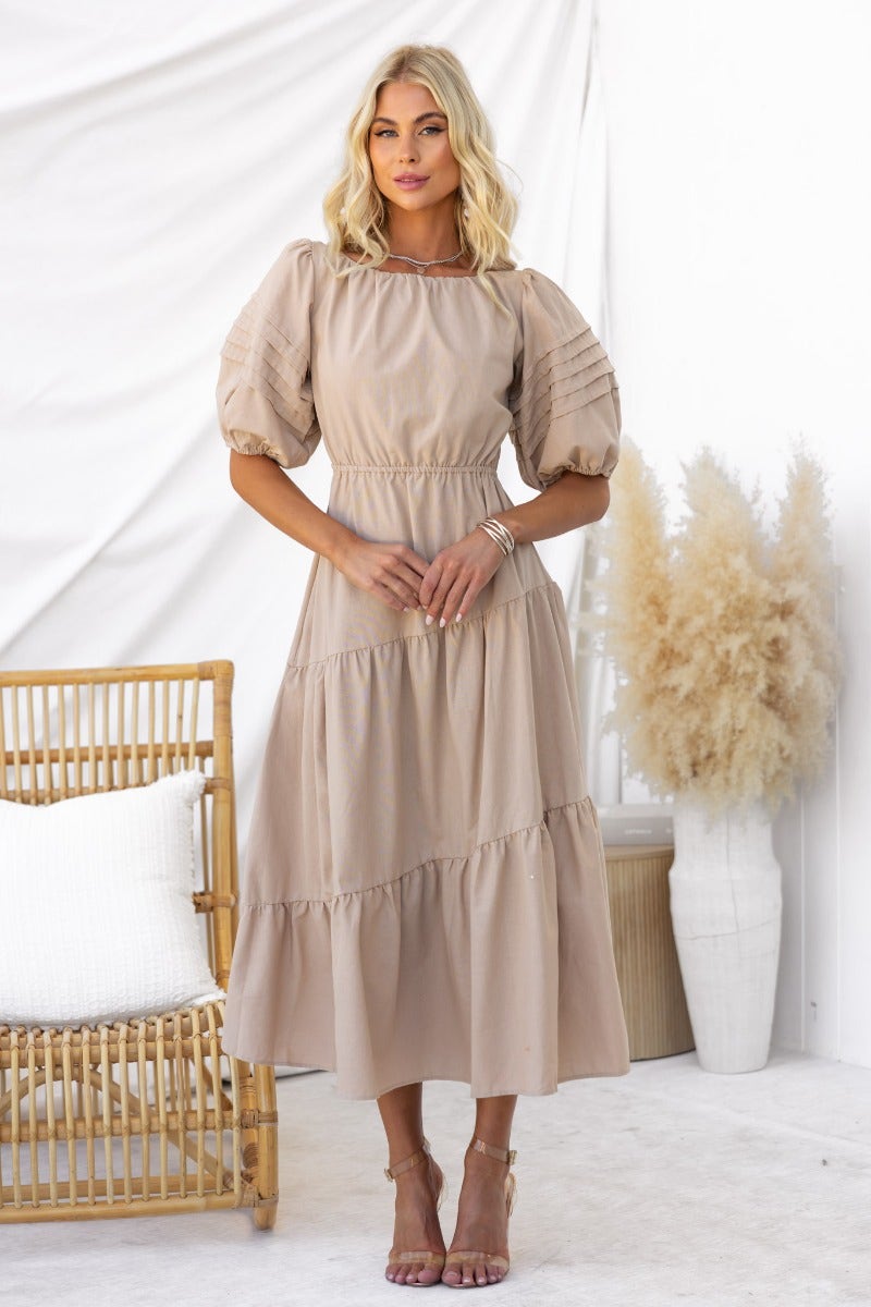 Oteo Midi Dress in beige with puff sleeves, a tie back, and tiered skirt, worn by a model in a styled setting.