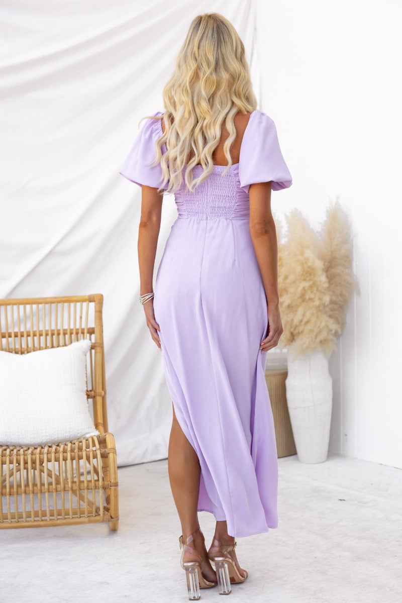 Back view of a woman in a purple maxi dress with square neckline and elastic sleeves, showcasing an asymmetric hem.