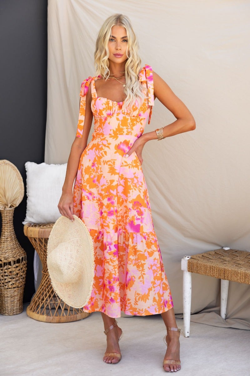 Model wearing a floral print Gratified midi dress with adjustable straps, holding a hat, against a light backdrop.