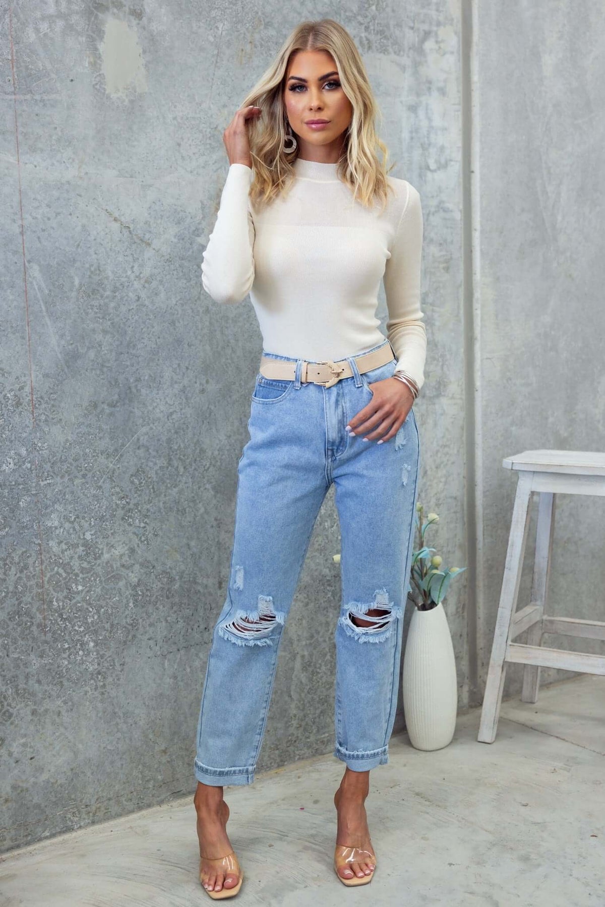 Model wearing a cream high-neck Milan Top paired with distressed blue jeans and sandals, showcasing a stylish look.