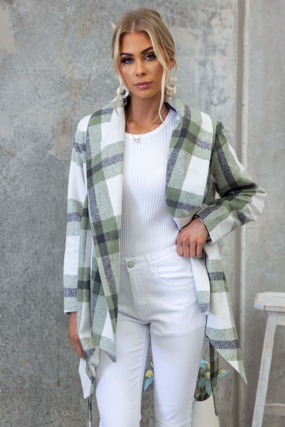 Stylish woman wearing a Phillips coat with plaid design over a white top and jeans, posing against a textured background.