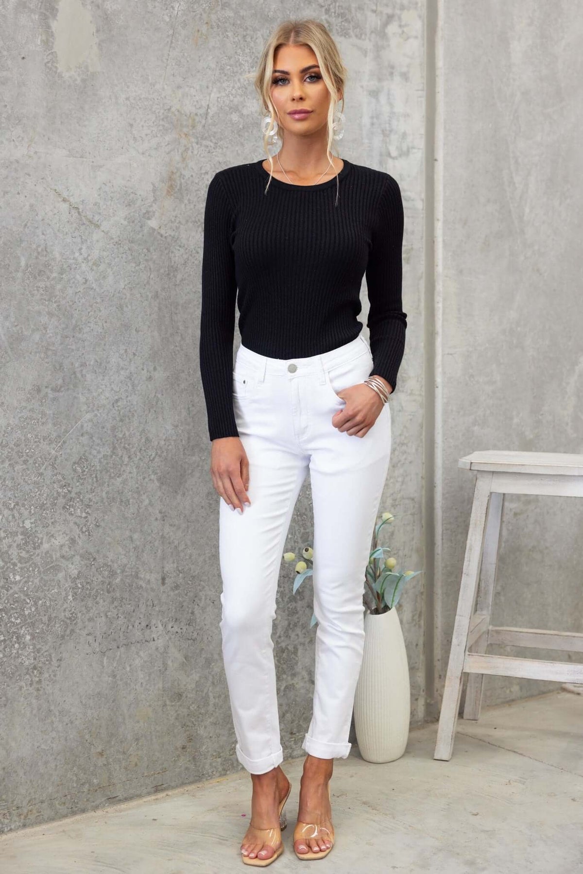Woman in a black ribbed top and white jeans standing against a concrete wall, showcasing a stylish casual look.