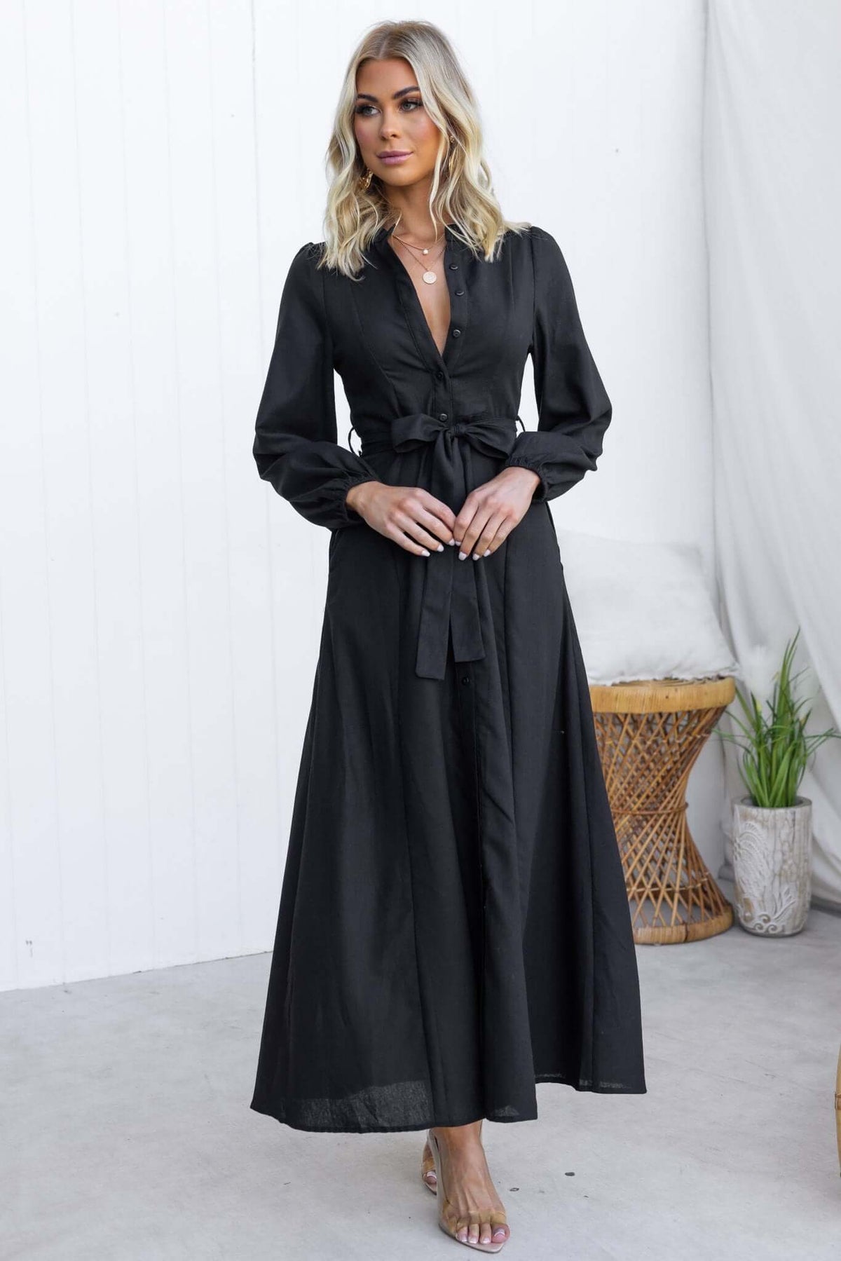 Model wearing a black Delgado Maxi Dress with a button-up front, elastic sleeves, and removable tie.