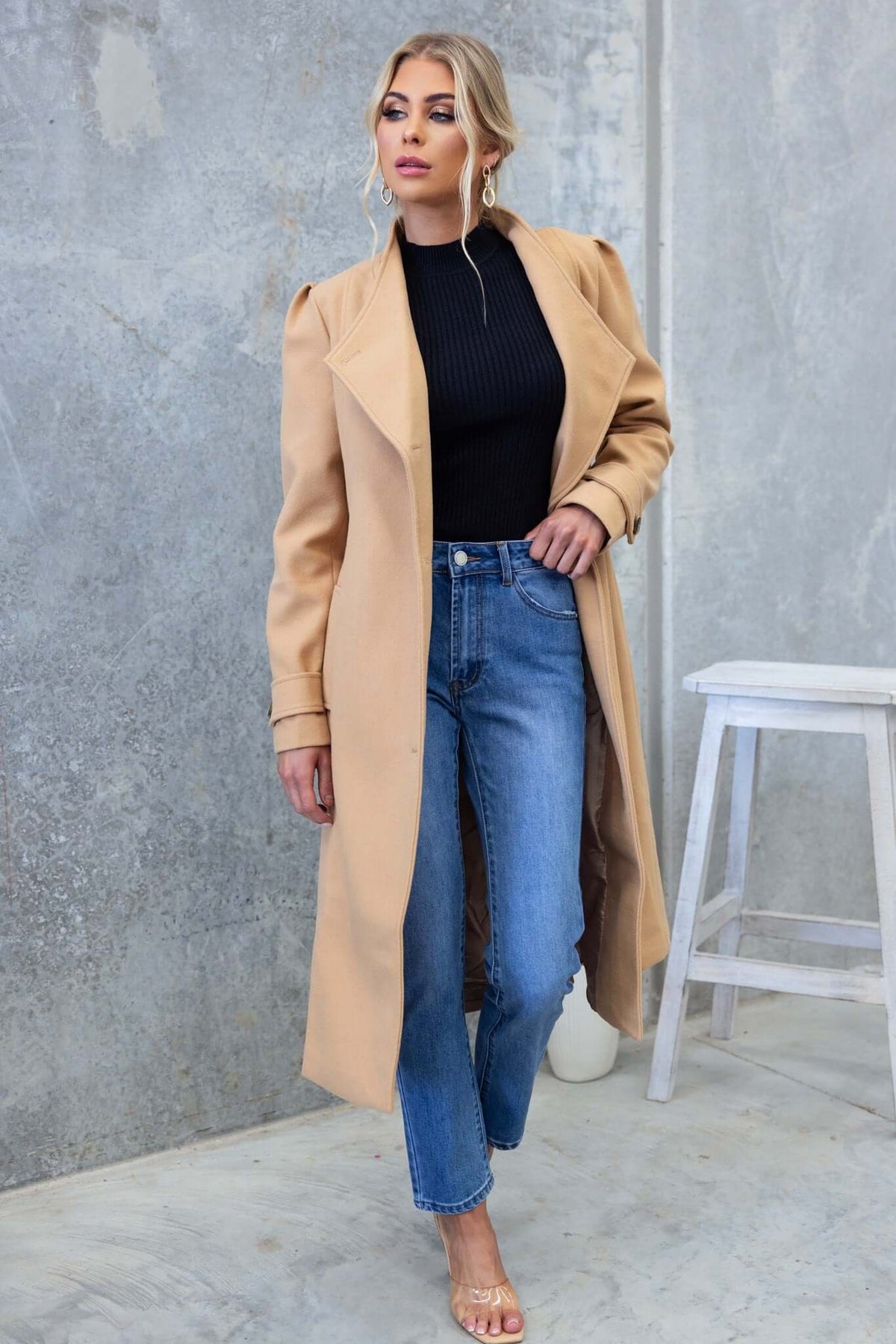 Model wearing a longline Ortona coat with a collar, paired with a black top and blue jeans against a textured backdrop.