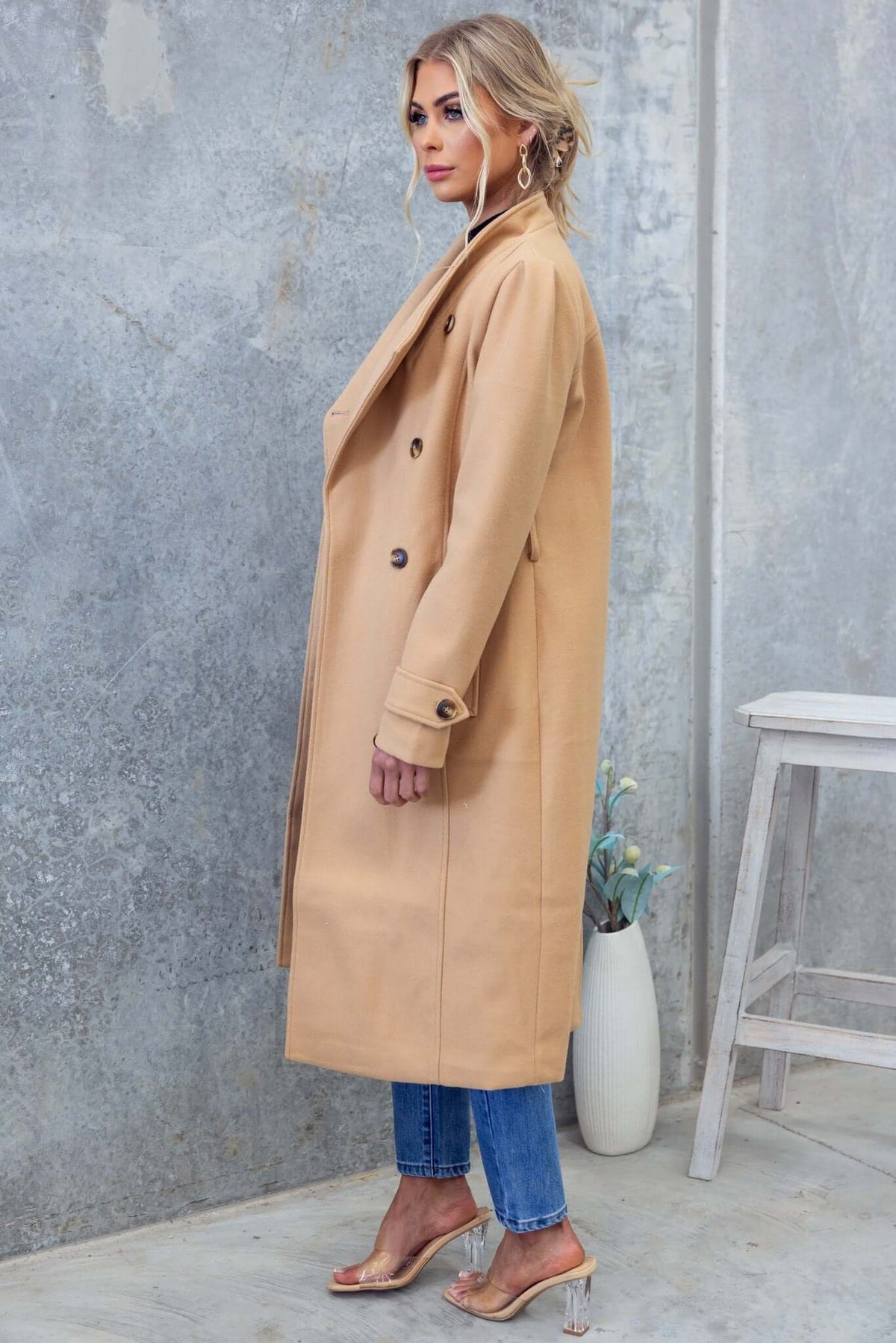 Side view of a model wearing a beige longline Ortona Coat with collar and lapel, paired with blue jeans.