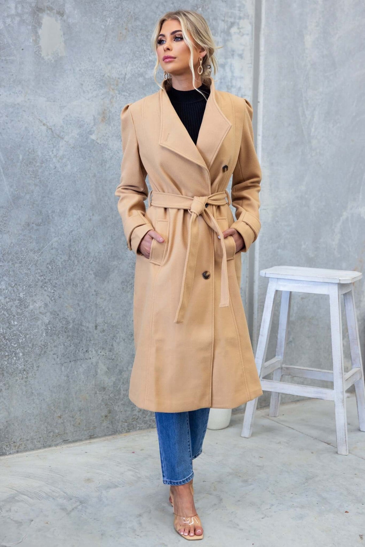 Model wearing a longline camel Ortona coat with a collar, lapel, and removable tie, styled over blue jeans.